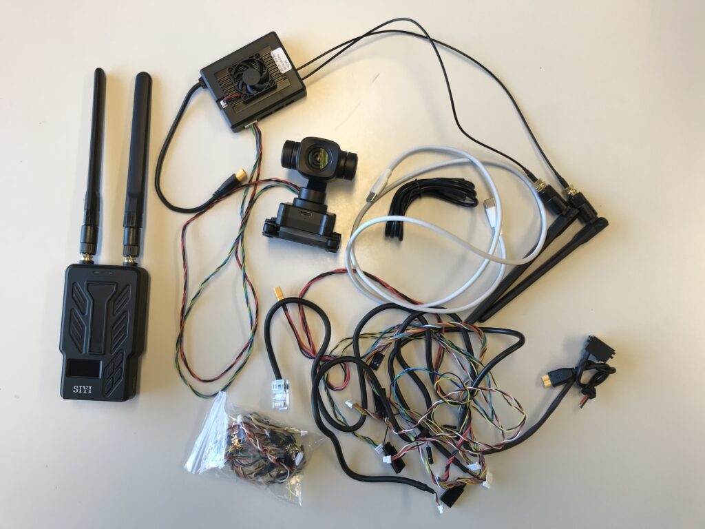 Yhteiskuvassa dronen kamera, videolähetin ja vastaanotin, sekä 15 kappaleen nippu erilaisia kytkentäjohtoja.