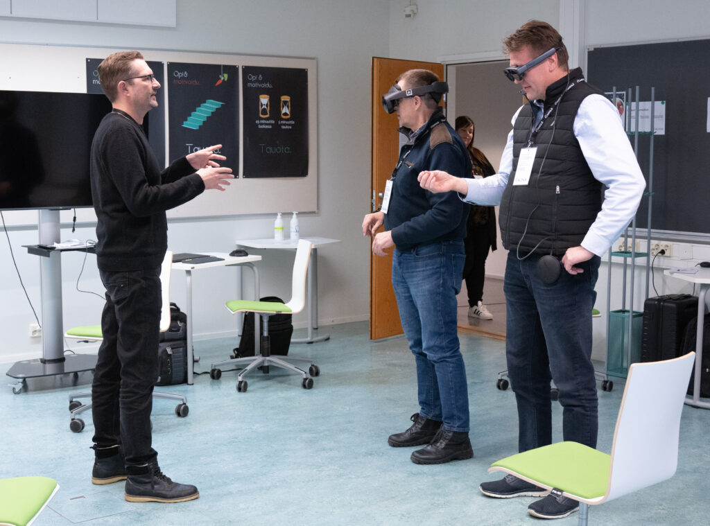 Anarky Labsin toimitusjohtaja ohjaa luokkahuoneessa kahta loppukäyttäjien edustajaa virtuaaliratkaisun tarkastelussa. Loppukäyttäjillä on VR-lasit päässä ja he ohjaavat saamiensa ohjeiden avulla kädet ilmassa omaa virtuaalinäkymäänsä.
