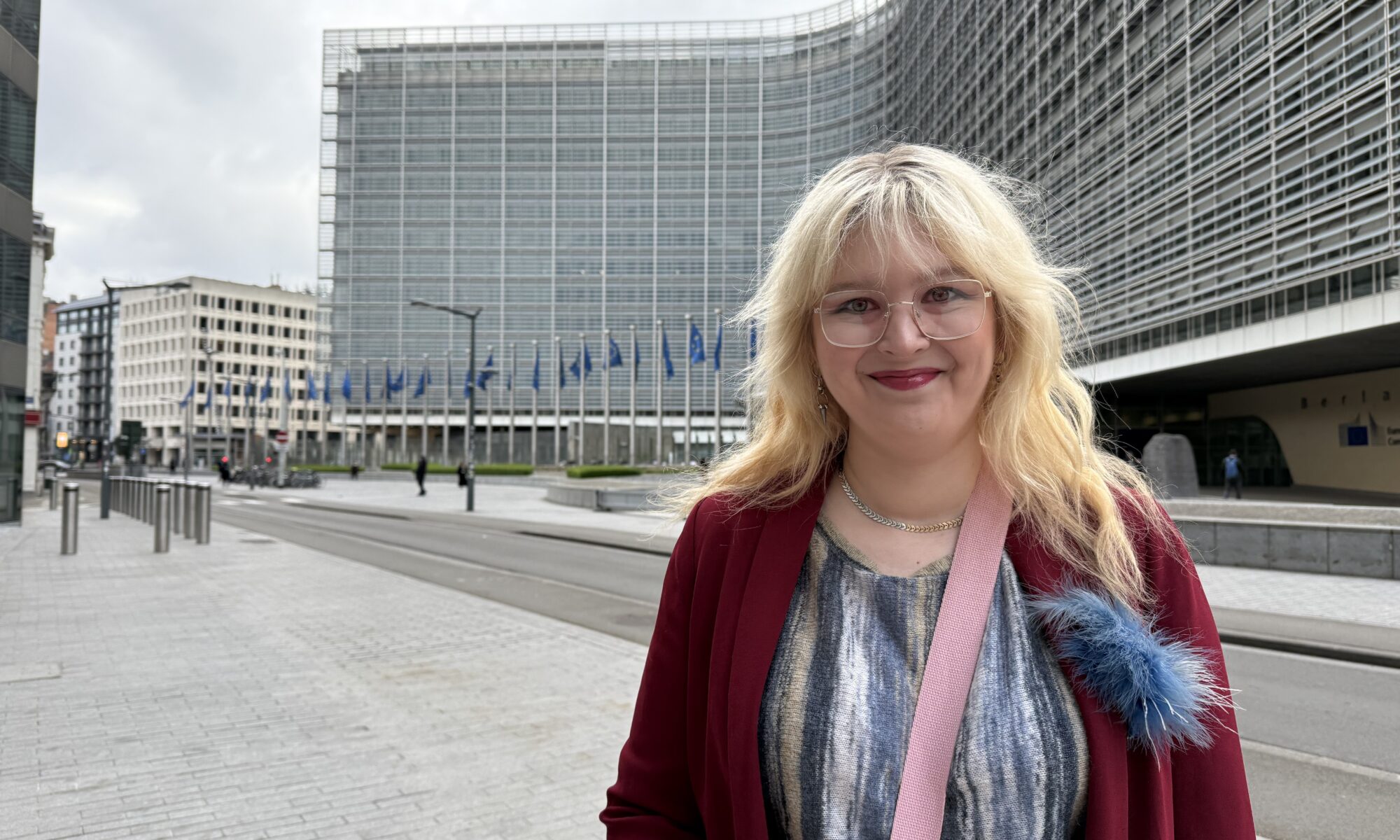 Leena Toivanen hymyilee kuvassa punainen samettitakki päällään. Hän seisoo Brysselissä EU-lippujen liehuessa taustalla.