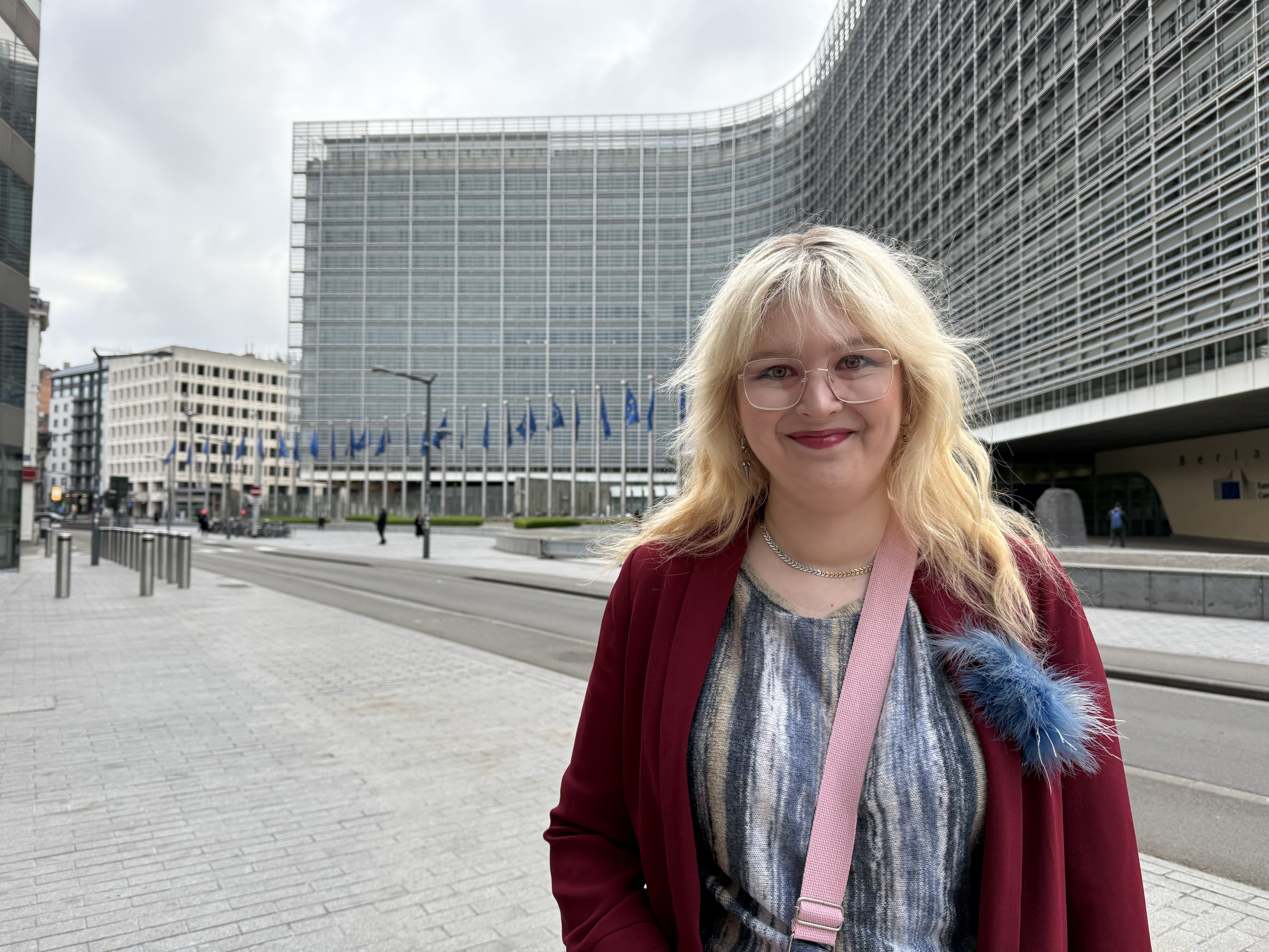 Leena Toivanen hymyilee kuvassa punainen samettitakki päällään. Hän seisoo Brysselissä EU-lippujen liehuessa taustalla.
