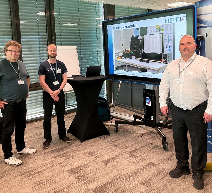 Kolme hankkeen osallistujaa seisoo esitysnäytön vieressä seminaaritilassa. Näytöllä esitellään LoLiPoP-IoT-hankkeen UC8-toteutusta ja lämmitysjärjestelmään liittyvää testausympäristöä.